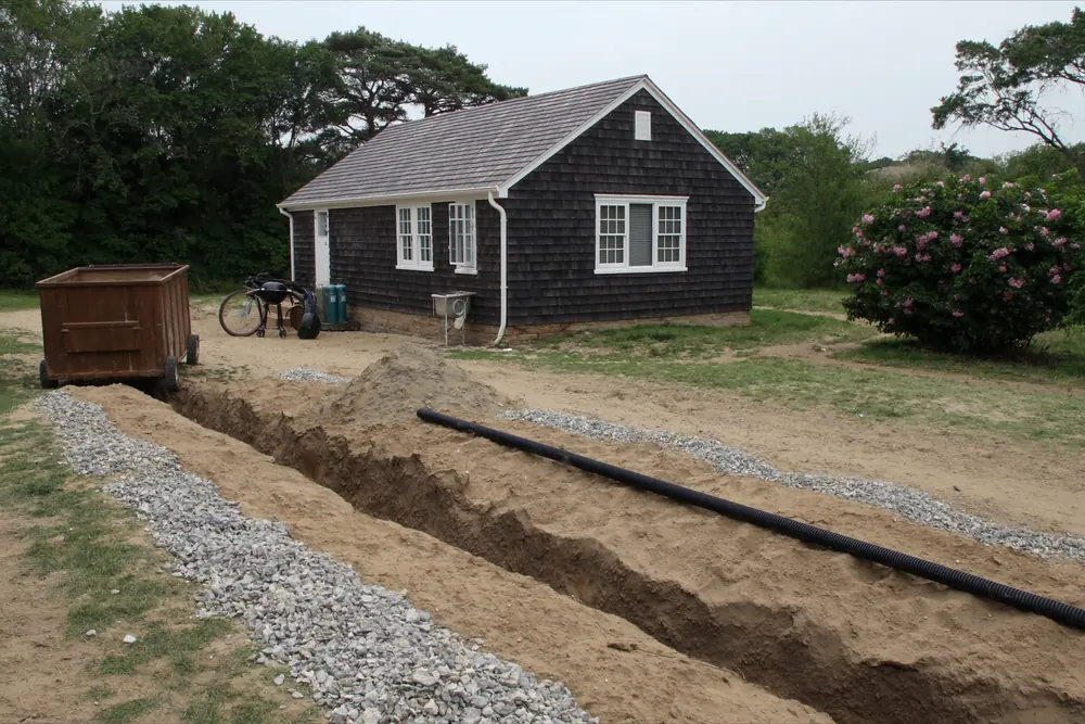 Septic leach field trenches filled with crushed stone and perforated distribution pipe dug in sandy soil behind a cedar shingled house in Barnstable, Massachusetts